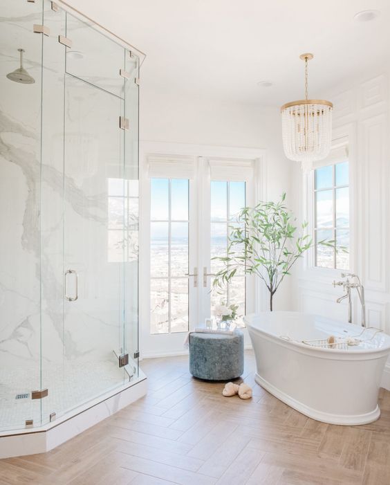 a neutral sophisticated bathroom with a dose of luxury - a white marble shower space, a parquet floor, an oval tub, a crystal chandelier and windows and a door to a terrace
