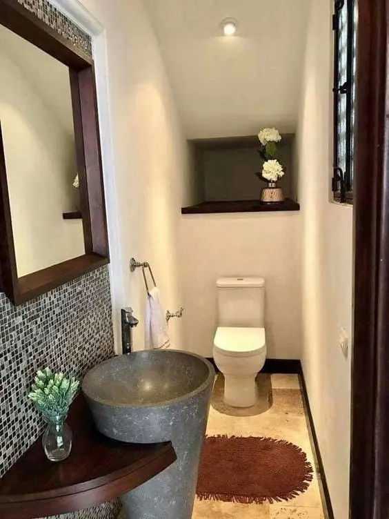 a powder room under the stairs with a mosaic tile wall, a free-standing stone sink, a toilet and a mirror in a dark-stained frame