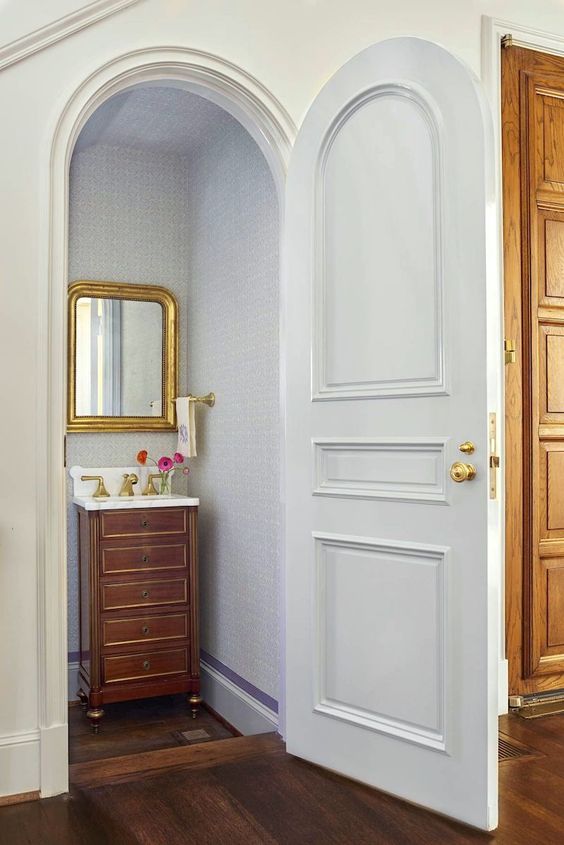 a refined powder room with an arched door, a stained vanity with drawers, a mirror in a gold frame under the stairs