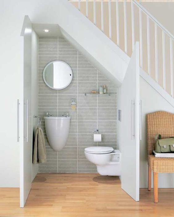 a simple and modern under stairs powder room with white floating appliances, a round mirror, a small shelf and some towels