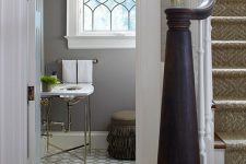 a small and elegant powder room with grey walls, a window, tiles on the floor, a vintage vanity with a sink and a pouf