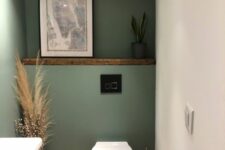 a small boho powder room under the stairs with green walls, a shelf with decor, a toilet, a vanity and some pampas grass