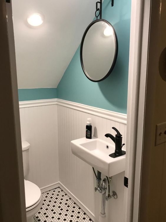 a small under the stairs powder room with turquoise walls and paneling, a printed floor, a wall-mounted sink, a hanging round mirror