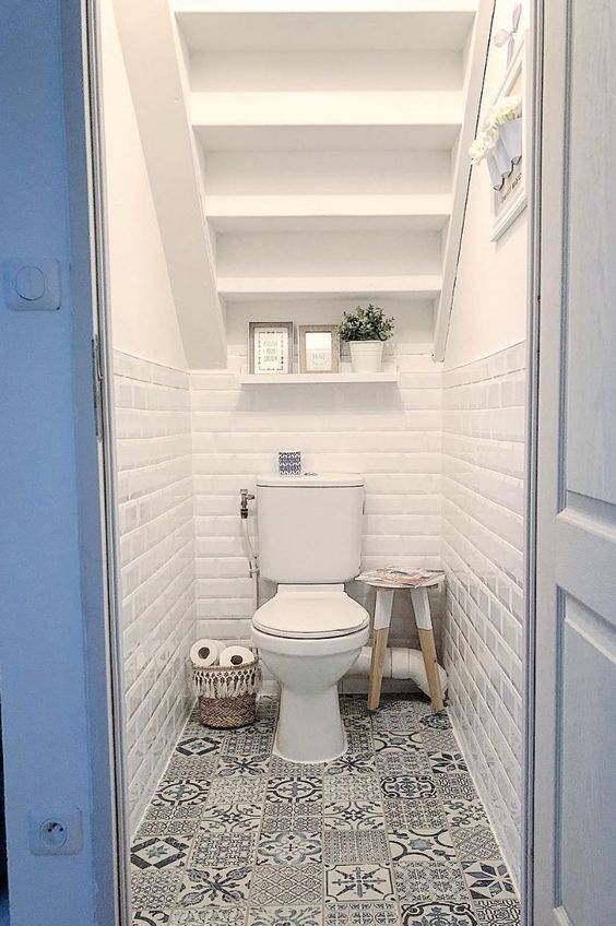 a small under the stairs powder room with white subway and printed tiles, an open shelf, some decor and a stool