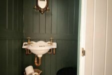a small vintage under the stairs powder room with dark green walls, a vintagw wall-mounted sink, a toilet, a basket and a tiny mirror