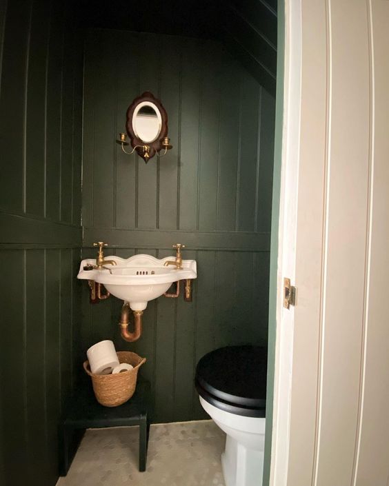 a small vintage under the stairs powder room with dark green walls, a vintagw wall-mounted sink, a toilet, a basket and a tiny mirror