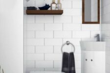 a small white powder room under the stairs clad with white subway tiles, white appliances, a wooden shelf and a mirror in a stained frame