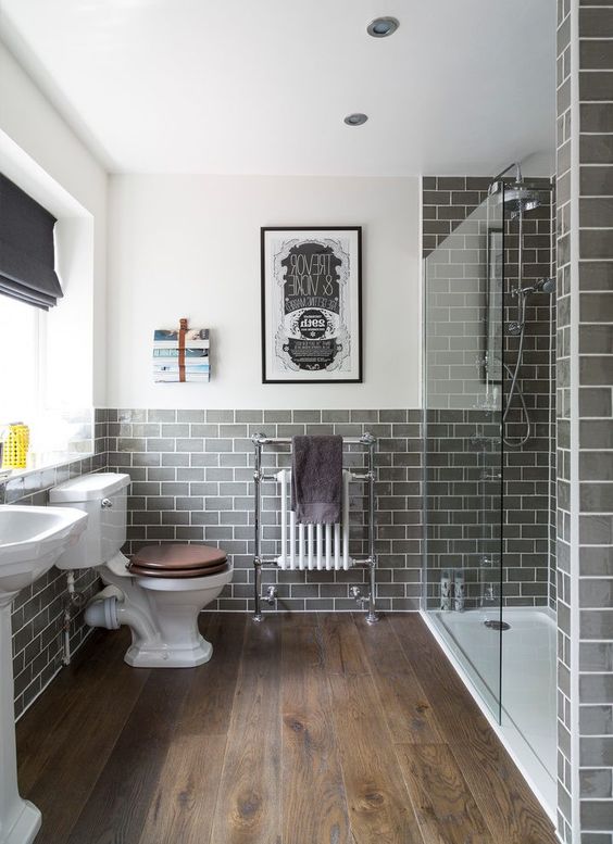 a stylish bathroom done with grey subway tiles and a rich-stained wooden floor, a large shower space enclosed in glass and vintage appliances