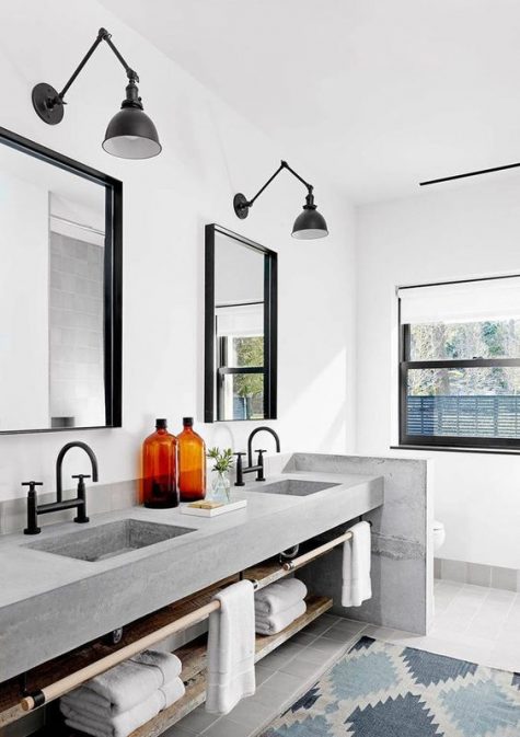 a stylish farmhouse bathroom with a large double vanity of concrete, two mirrors and black sconces plus a concrete space divider
