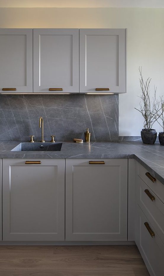 a stylish monochromatic grey kitchen with shaker style cabinets, a grey marble backsplash and countertops plus brass fixtures and handles