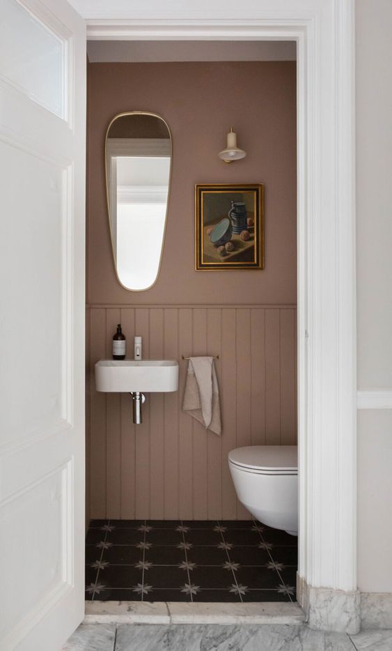 a tiny moody powder room under stairs, with mauve walls and wainscoting, white appliances and a catchy mirror is amazing