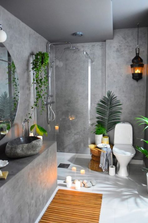 a tropical inspired bathroom of concrete, with a sleek vanity, a wooden bath mat and a stool, potted plants and lots of candles