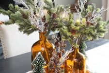 a woodland Christmas centerpiece of evergreens and pinecones in amber bottles, bottlebrush trees and lights