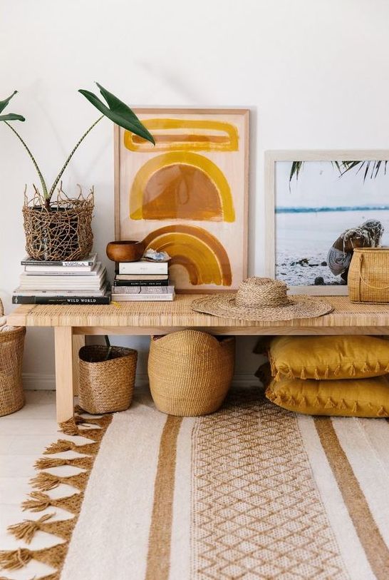 a bright boho space with a woven bench, mustard-colored pillows, bright artwork, baskets and a potted plant plus a cool rug