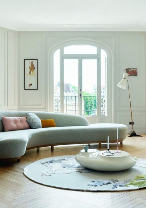 a light filled living room with a grey curved sofa, a unique round coffee table, a printed round rug and a chic floor lamp