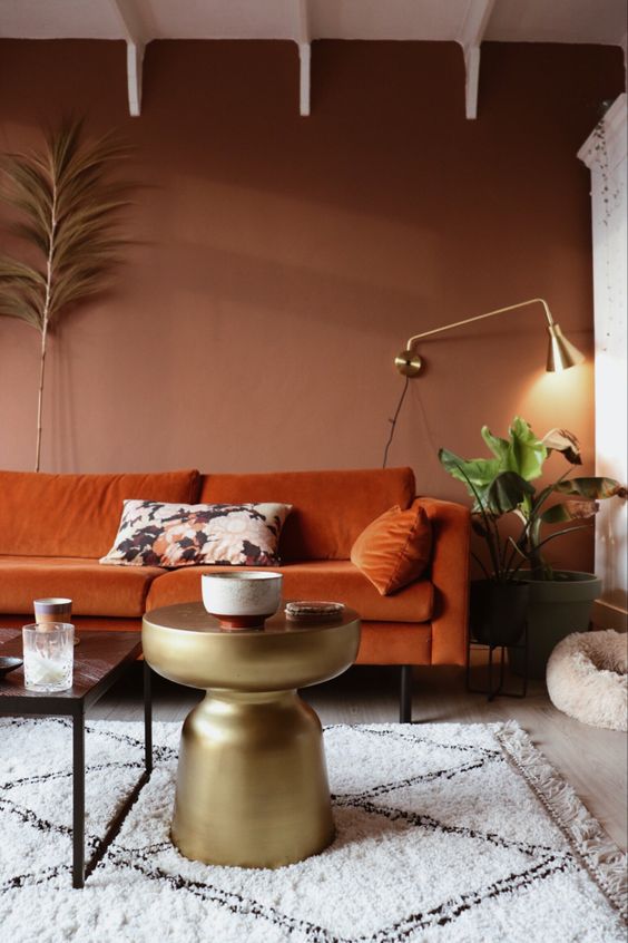 a beautiful earthy tone living room with brown walls, a rust colored sofa, a gold side table and a gold sconce and potted greenery