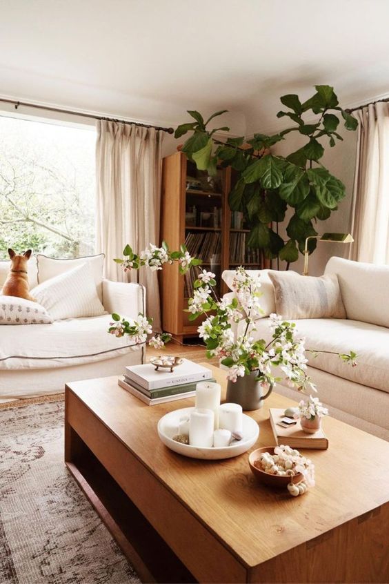 a warm neutral living room done in soft creamy shades, with light-stained furniture, potted plants and blooms and a printed rug