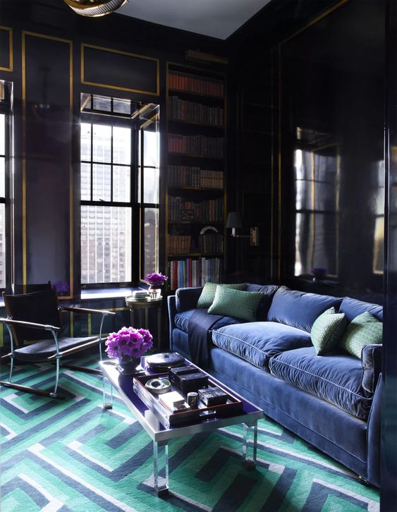 a moody living room with deep purple walls, touches of gold, built in bookcases, a very peri velvet sofa and a green and very peri geo rug