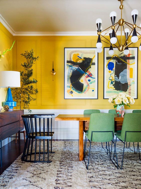 a modern boho dining room with sunny yellow panels, a stained table, green chairs, a brass, black chandelier and greenery