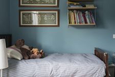 a stone blue kid’s room with a bookshelf, a stained bed, some art and a basket with a lid for storage
