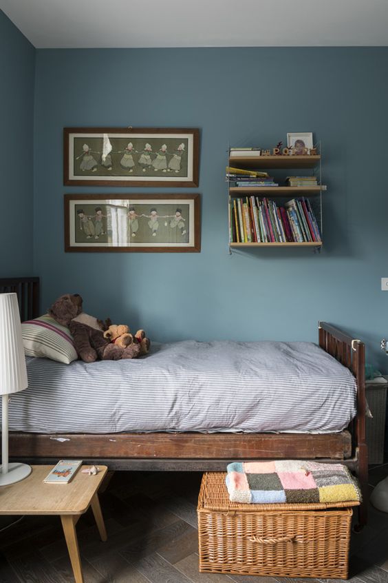 a stone blue kid's room with a bookshelf, a stained bed, some art and a basket with a lid for storage