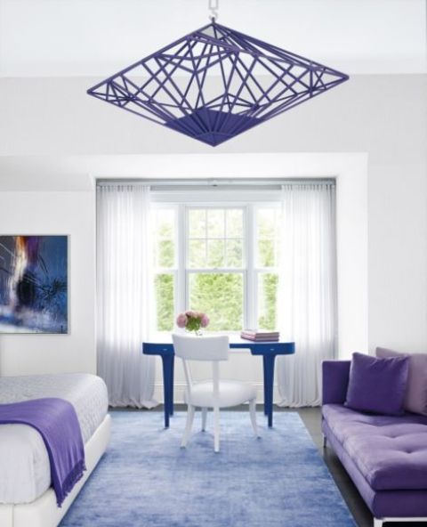 a neutral space with a white bed with a very peri blanket, a very peri daybed, a bold violet chandelier and a blue desk