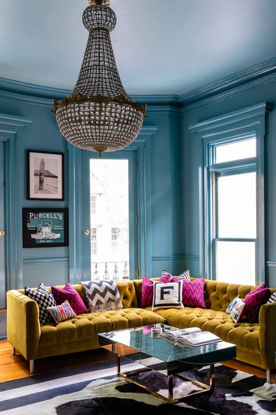 a super bold and refined living room with stone blue walls, a mustard sectional sofa, pink pillows and a statement chandelier