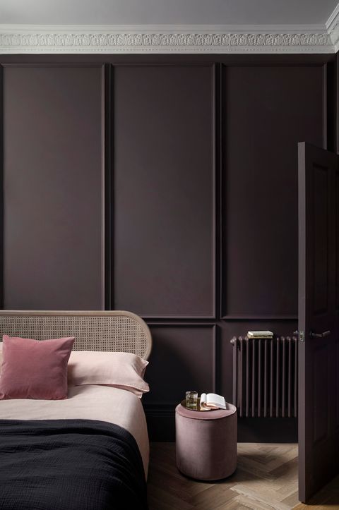 a minimalist bedroom with deep purple walls, a door and a radiator, a bed with a cane headboard and a pink stool