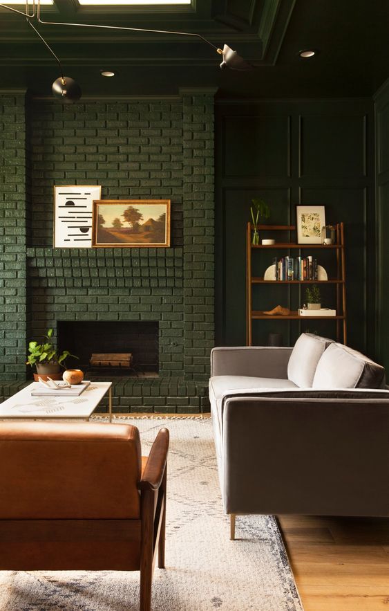 a moody and elegant living room with dark green walls and a dark green brick fireplace, an amber leather chair, a grey sofa and brass lamps