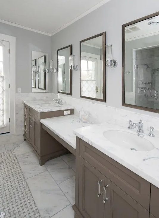 a beautiful bathroom clad with white stone tiles, a large taupe double vanity, mirrors in brown frames, sconces and vintage fixtures