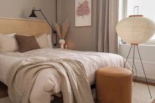 a boho taupe bedroom with a rattan bed, neutral bedding, an orange leather stool, a floor lamp and some art and pampas grass