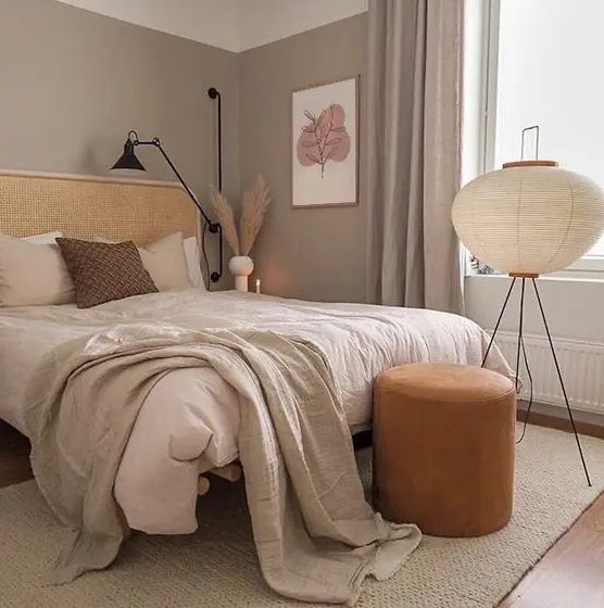 a boho taupe bedroom with a rattan bed, neutral bedding, an orange leather stool, a floor lamp and some art and pampas grass