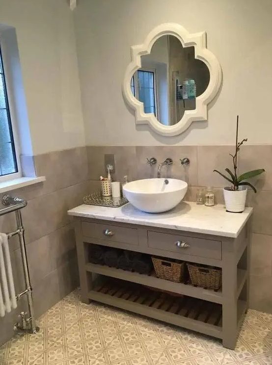 a chic bathroom with taupe and patterned tiles, a taupe vanity, a mirror in a refined frame, a bowl sink and a potted bloom is cool