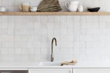 a clean white kitchen with plain cabinetry and white stone countertops, a white Zellige tile backsplash and light-stained open shelves