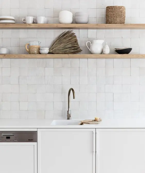 a clean white kitchen with plain cabinetry and white stone countertops, a white Zellige tile backsplash and light-stained open shelves