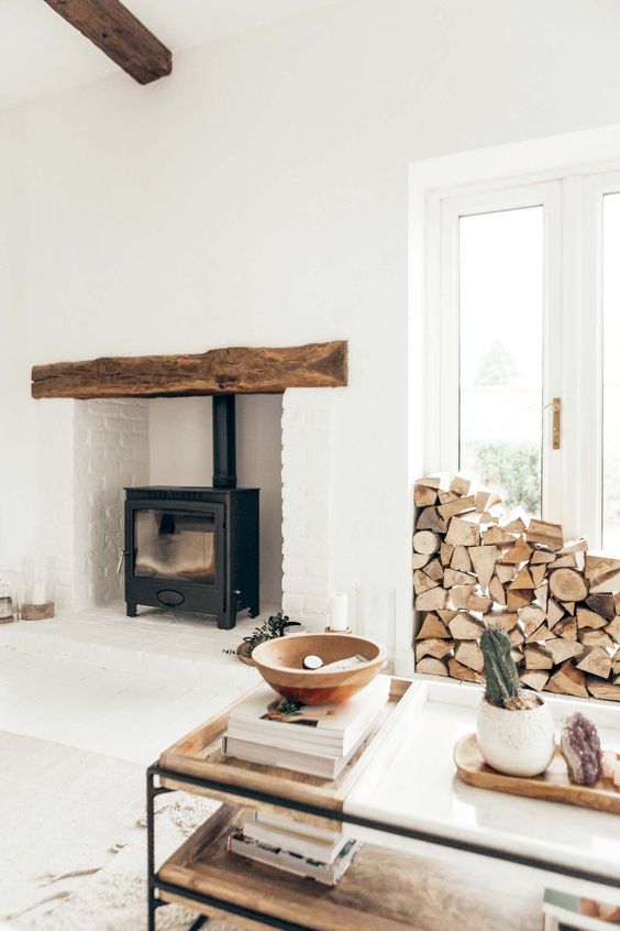 a cozy farmhouse space with white birck and a black hearth, a wood slab mantel, firewood storage by the doors to the garden