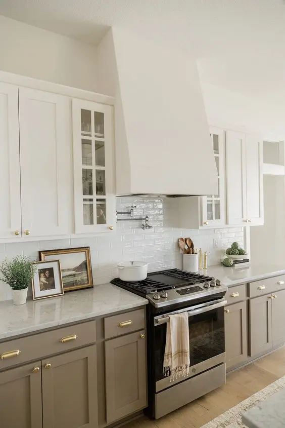 a delicate kitchen with white upper cabinets and taupe lower ones, a white tile backsplash, gold knobs for a chic and elegant touch