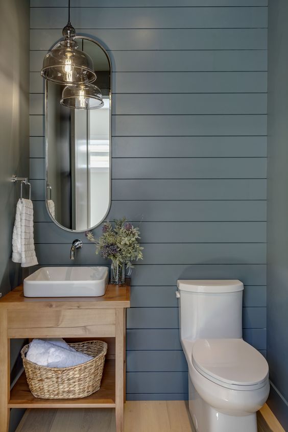 a farmhouse powder room with slate grey shiplap walls, a small stained vanity, a catchy curved mirror and a pendant lamp