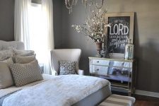a glam bedroom with taupe walls, an upholstered bed, neutral bedding, a striped bench, a creamy chair, a vintage chandelier