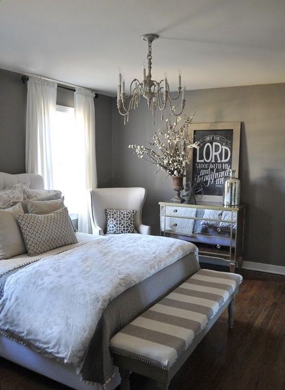 a glam bedroom with taupe walls, an upholstered bed, neutral bedding, a striped bench, a creamy chair, a vintage chandelier