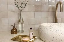 a gorgeous bathroom with a vintage feel, with neutral Zellige tiles, a terrazzo sink, a white stone countertop and brass fixtures