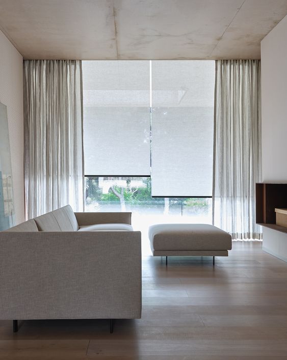 a minimalist living room done in neutrals, with classic blinds and semi-sheer neutral curtains to get privacy yet not to block out excessive sunshine