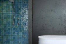 a modern bathroom with a dark concrete part with a white metal tub and a shower space clad with green and blue Zellige tiles