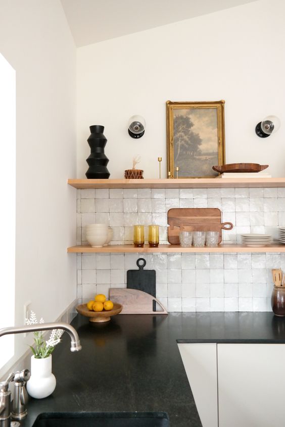 a modern contrasting kitchen with sleek white cabinets, black countertops, a white Zellige tile backsplash and open shelves