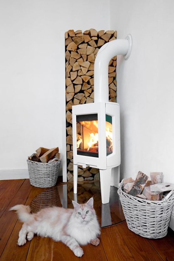 a modern white wood burning stove on a glass stand, with firewood in the corner and baskets for firewood
