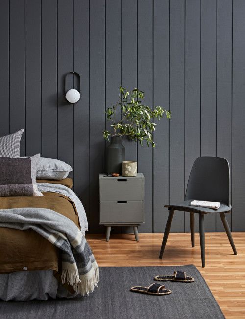 a moody bedroom with black shiplap walls, a bed with pretty bedding, a grey nightstand, a grey chair and greenery branches