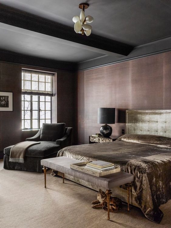 a moody bedroom with taupe grasscloth wallpaper, a black ceiling and beams, a gold velvet bed, a taupe velvet bench and a black lounger