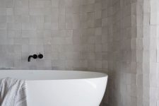a refined neutral bathroom clad with neutral Zellige tiles all over, an oval tub and black fixtures for a super modern feel