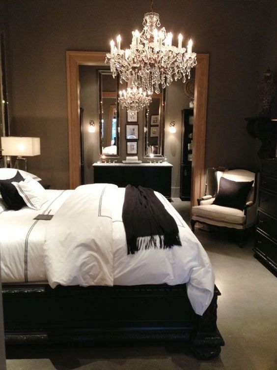a refined taupe bedroom with a black bed and black and white bedding, a black dresser, white chairs and a crystal chandelier