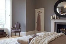 a refined taupe bedroom with molding, a neutral bed and bench, cozy blankets, a chic chair and a vintage fireplace
